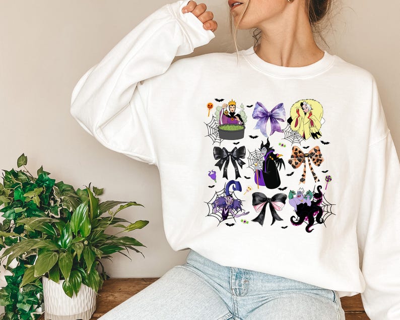 K&ouml;nnte beinhalten: Wei&szlig;es Sweatshirt mit einem farbenfrohen Grafikdesign mit Halloween-Motiven, darunter Schleifen und Charaktere. Das Design zeigt eine Hexe, eine K&ouml;nigin und andere gruselige Elemente. Das Design ist auf der Vorderseite des lang&auml;rmeligen Oberteils aufgedruckt.