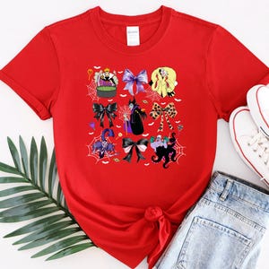 K&ouml;nnte beinhalten: Ein rotes T-Shirt mit einem farbenfrohen Grafikdesign verschiedener Cartoon-Charaktere und Schleifen. Das Design enth&auml;lt Bilder einer Hexe, eines Kessels und Spinnweben. Das Shirt wird mit blauen Jeans und wei&szlig;en Turnschuhen kombiniert.