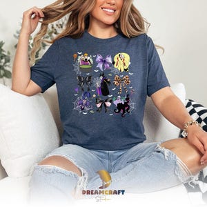 K&ouml;nnte beinhalten: Ein marineblaues T-Shirt mit einem farbenfrohen Grafikdesign von Halloween-Charakteren und Schleifen. Das Design enth&auml;lt Bilder von Hexen, Spinnen und anderen gruseligen Elementen. Der Text "DREAMCRAFT Studio" ist unten sichtbar.