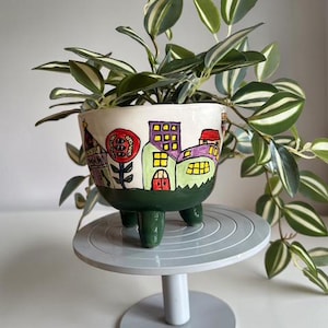 Op de afbeelding: Een handbeschilderde keramische plantenpot met een grillig stadsgezichtontwerp. De witte pot heeft kleurrijke gebouwen, een rode zon en groene accenten. De pot staat op drie groene poten en bevat een hangplant met groen-wit gestreepte bladeren.