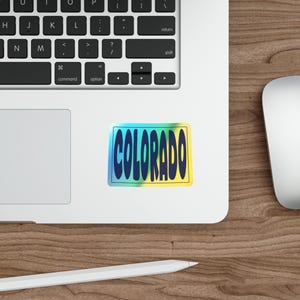 May include: A rectangular sticker with the word "COLORADO" in bold, blue letters. The letters have a slight shadow effect. The background transitions from blue to yellow, with a black border. The sticker is on a white surface.