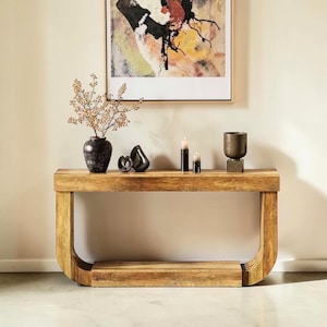 Peut inclure: Une console en bois avec un design incurvé, présentant une finition en bois naturel. La table est ornée d'objets décoratifs, dont un vase avec des branches séchées, des sculptures noires et des bougies. Une peinture abstraite est accrochée au mur.