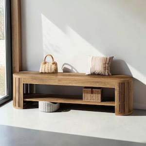 May include: A light brown wooden bench with a curved design, featuring a lower shelf. A woven handbag, a striped cushion, and two woven baskets are placed on the bench. The bench is in a room with a window.