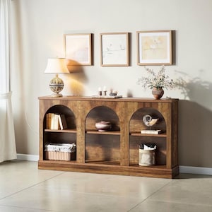 May include: A wooden bookshelf with three arched sections, each with a shelf. Books, decorative items, and baskets are displayed. A lamp and framed artwork are above the unit. The wood has a warm, natural finish.