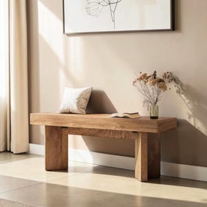 May include: A wooden bench with a rectangular top and two thick, rectangular legs. A white cushion sits on the bench, alongside an open book and a small vase of dried flowers. The bench is made of light brown wood.