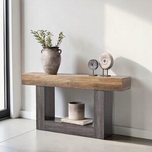 May include: A contemporary wooden console table with a light brown top and grey legs. It features a vase of greenery, two stone wheel ornaments, a small ceramic pot, and a book. This console table is a stylish addition to any living space.