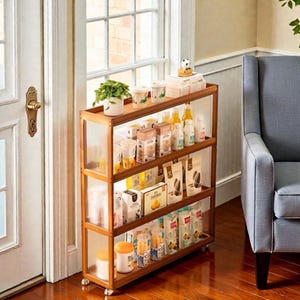 May include: A three-tiered wooden storage shelf with clear, textured side panels. The shelves are filled with various packaged food items, drinks, and a small potted plant. The shelf is on wheels for easy movement.