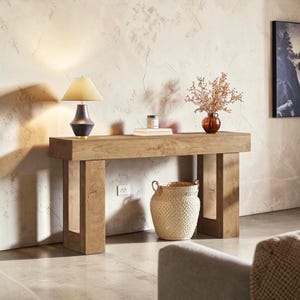 May include: A light brown wooden console table with a rectangular top and four sturdy legs. A dark lamp with a beige shade sits on the left side. A woven basket and a small vase with dried flowers are also on the table.