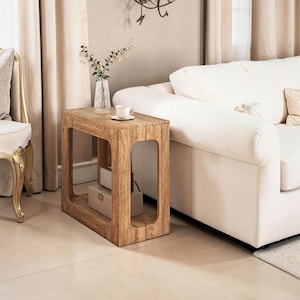 May include: A wooden side table with a natural finish. The table has a rectangular shape with cut-out sides and an internal shelf. A white teacup and saucer are on top, with a vase of flowers. Home furniture.