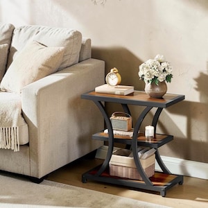 May include: A three-tier side table featuring a black metal frame with curved supports and rectangular wooden shelves. The top shelf holds a gold alarm clock, a book, and a vase of white flowers. The middle shelf holds a small radio and a candle.