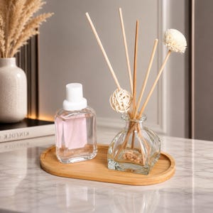 May include: A clear glass reed diffuser with wooden sticks, a decorative woven sphere, and a dried flower, paired with a clear glass bottle with a white cap on an oval bamboo tray. The diffuser contains small light brown stones at the base.