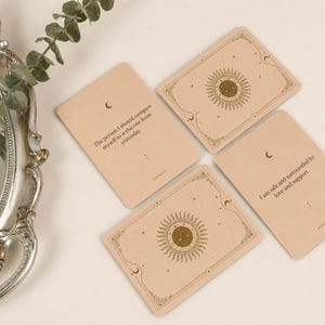 May include: Four beige oracle cards with celestial designs and text. Each card features a sunburst design, crescent moons, and decorative borders. The cards have phrases like "The person I should compare myself to is the one from yesterday."