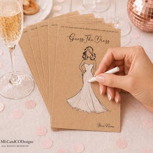 May include: A pile of kraft paper bridal shower game cards with the text "Guess The Dress" and a drawing of a wedding dress. A hand is holding a white pen, ready to write. A glass of champagne is in the foreground.