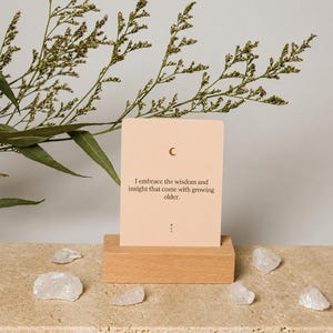 May include: A beige card with a crescent moon symbol and the text "I embrace the wisdom and insight that come with growing older." is displayed on a wooden block. Clear quartz crystals are scattered around the base.