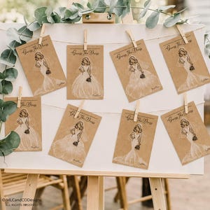 May include: An easel holds eight cards with illustrations of wedding dresses, clipped to a string. Each card, made of kraft paper, has the text "Guess The Dress". The cards are part of a bridal shower game.