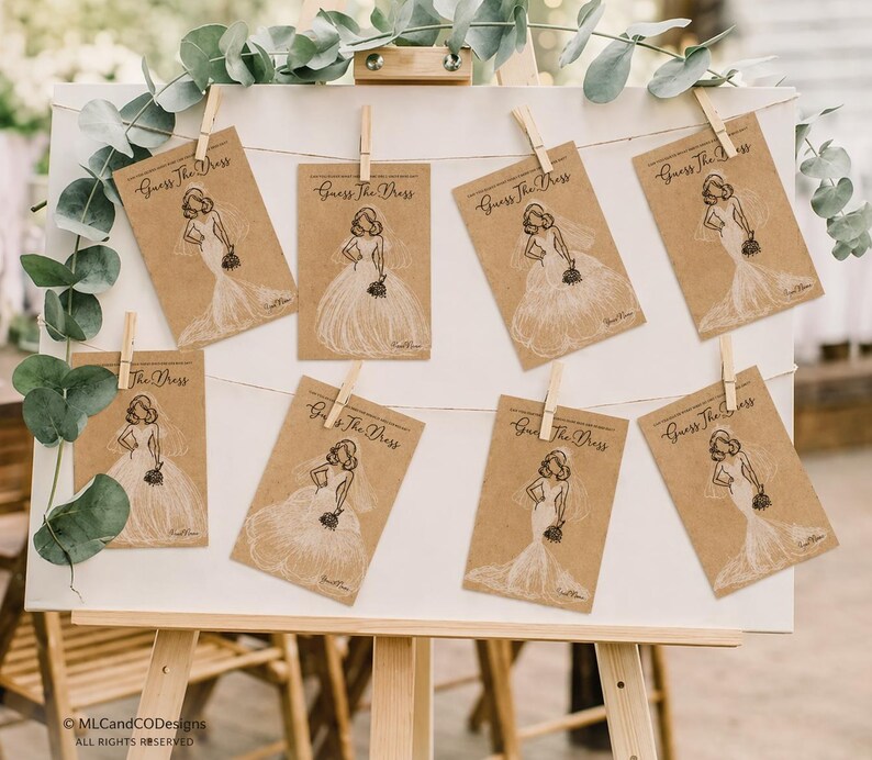 May include: An easel holds eight cards with illustrations of wedding dresses, clipped to a string. Each card, made of kraft paper, has the text "Guess The Dress". The cards are part of a bridal shower game.