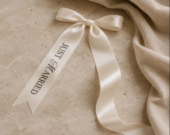May include: A cream-coloured satin ribbon tied into a bow with two long tails. The left tail features the black printed text "JUST MARRIED" in a serif font. The ribbon has a smooth, reflective finish and ends in a V-shaped cut on the left side.