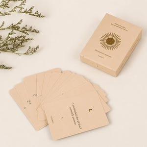 May include: A beige affirmation card deck with a matching box. The box features a sunburst design and the words "Affirmations Moments" and "Embrace Your Spirit, Brighten and Follow Your Star." Several cards are fanned out, with text including "I am thankful for all that I currently possess."