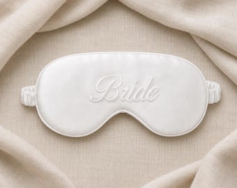 May include: A white sleep mask featuring the word Bride embroidered in a matching white script font. The mask has a smooth, satin-like finish and includes a gathered elastic strap on each side for a secure fit around the head.