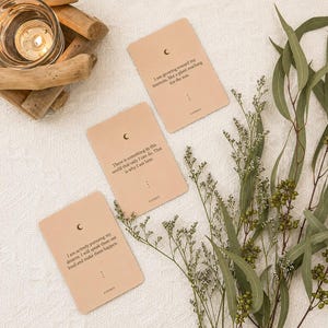 May include: Three beige affirmation cards with a crescent moon symbol and text. The cards are surrounded by greenery and a lit candle in a glass holder. The cards have inspirational quotes.