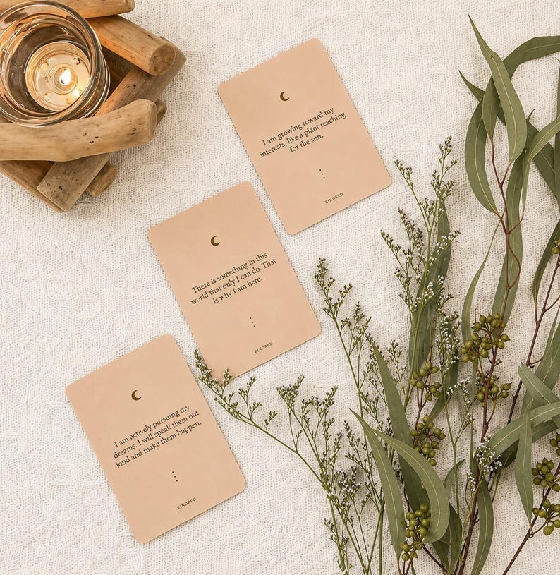 May include: Three beige affirmation cards with a crescent moon symbol and text. The cards are surrounded by greenery and a lit candle in a glass holder. The cards have inspirational quotes.