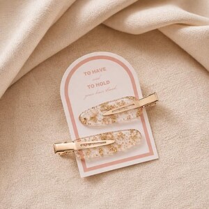 May include: Two hair clips with clear resin and gold leaf flakes attached to a white display card with a pink arch border. The card features the text TO HAVE and TO HOLD your hair back in a light brown font. The clips have gold metal hardware.