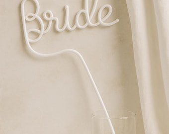 May include: A white, cursive "Bride" sign, possibly for a wedding. The sign is made of a continuous line and is positioned above a champagne flute with a white straw. The background is a neutral colour.