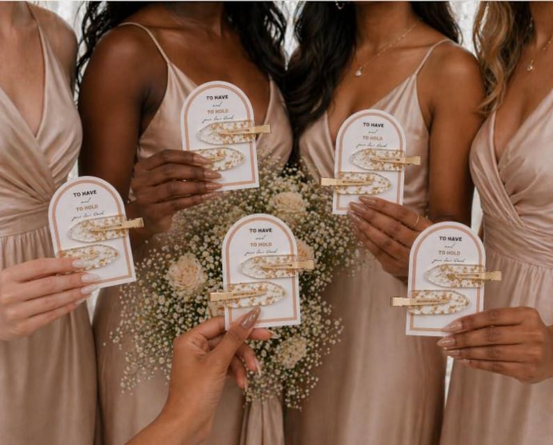May include: Five sets of two hair clips with gold-toned metal alligator clasps and white resin pieces containing gold flakes. Each set is attached to an arched white card featuring the text To Have and To Hold your hair back.