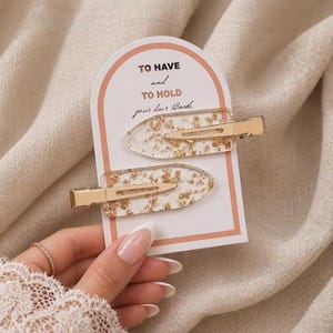 May include: Two clear resin hair clips with embedded gold leaf flakes, attached to a white arched display card featuring the text TO HAVE and TO HOLD your hair Back in black and peach lettering. Each clip has a gold-toned metal alligator clasp.