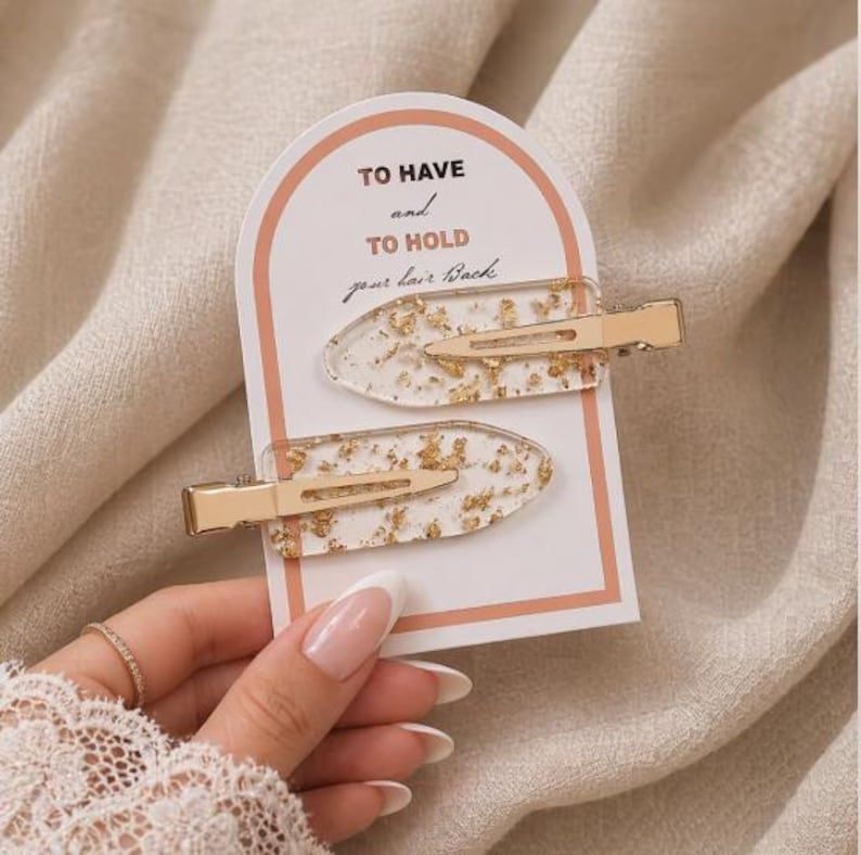May include: Two clear resin hair clips with embedded gold leaf flakes, attached to a white arched display card featuring the text TO HAVE and TO HOLD your hair Back in black and peach lettering. Each clip has a gold-toned metal alligator clasp.