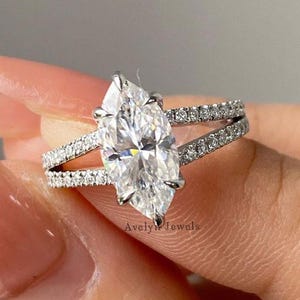 May include: A marquise-cut diamond engagement ring with a split band. The ring features a large, clear marquise-cut diamond as the centerpiece, with smaller diamonds set along the split band. The ring is set in a silver-colored metal.