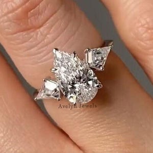 Pode incluir: Um close-up de um anel de diamante com uma pedra central em forma de pêra e duas pedras laterais triangulares menores. O anel é cravejado em uma banda de prata. O anel está no dedo de uma pessoa.