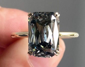 May include: A ring featuring a large, rectangular, faceted dark gray stone set in a four-prong gold setting. The stone displays deep charcoal tones with reflective surfaces, mounted on a thin, polished gold band that encircles the finger.