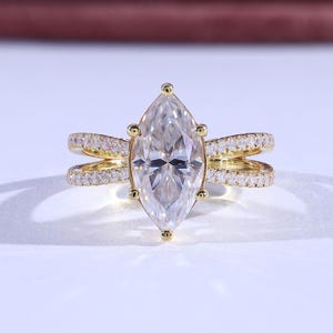 Pode incluir: Um anel de ouro com uma grande pedra preciosa transparente em corte marquesa como peça central. A banda divide-se em duas filas paralelas, cada uma incrustada com pedras preciosas transparentes menores. O anel está sobre uma superfície branca.