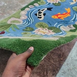 May include: A rectangular rug featuring a pond scene with koi fish, water lilies, and a green border. The rug's design includes a blue pond with white water lines, and green and beige accents. The rug is held up by a hand.