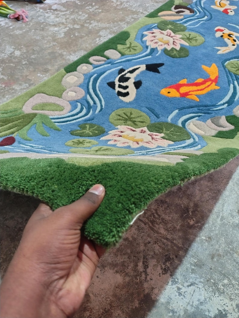 May include: A rectangular rug featuring a pond scene with koi fish, water lilies, and a green border. The rug's design includes a blue pond with white water lines, and green and beige accents. The rug is held up by a hand.
