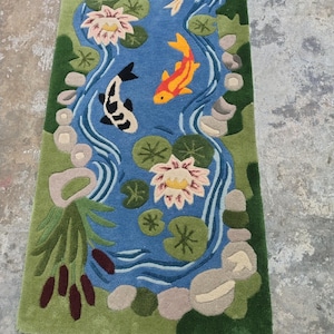 May include: A rectangular rug featuring a pond scene with a blue water background. The rug has green edges and depicts koi fish, lily pads, and water lilies in shades of orange, white, and pink. The rug is approximately 60 inches long.