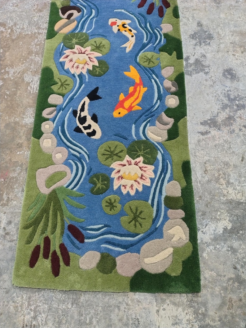May include: A rectangular rug featuring a pond scene with a blue water background. The rug has green edges and depicts koi fish, lily pads, and water lilies in shades of orange, white, and pink. The rug is approximately 60 inches long.