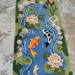 May include: A rectangular rug featuring a pond scene with koi fish, water lilies, and lily pads. The pond is blue, surrounded by green grass and rocks. The koi fish are orange, black, and white. The water lilies are pink and white.
