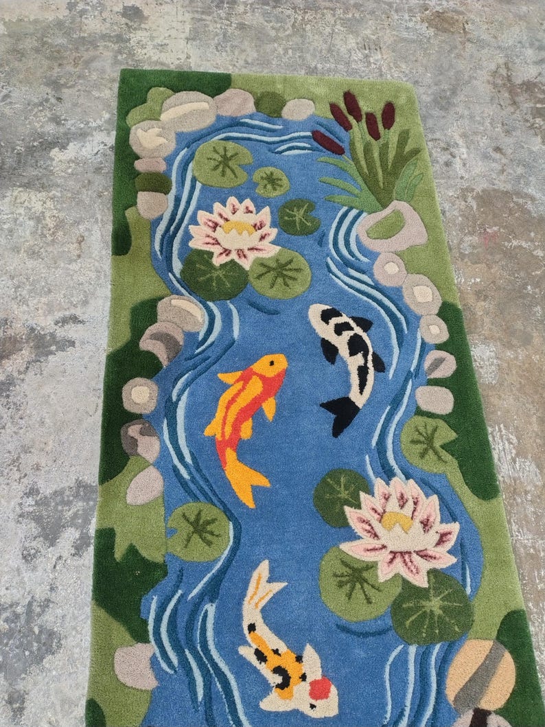 May include: A rectangular rug featuring a pond scene with koi fish, water lilies, and lily pads. The pond is blue, surrounded by green grass and rocks. The koi fish are orange, black, and white. The water lilies are pink and white.