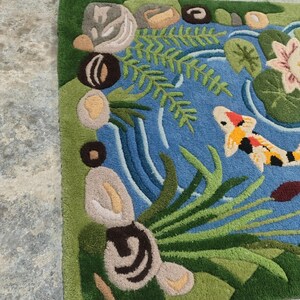 May include: A hand-tufted rug featuring a pond scene with a koi fish. The rug has a blue pond with green plants, lily pads, and a pink flower. The koi fish is orange, white, and black. The border is green.