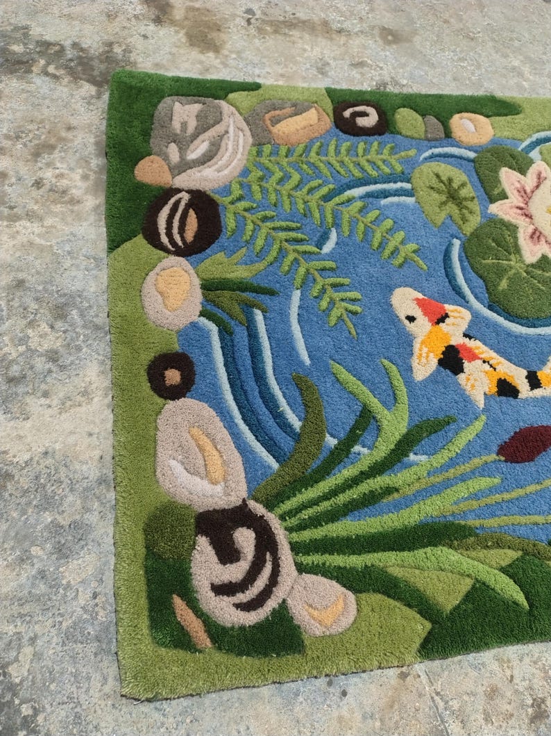May include: A hand-tufted rug featuring a pond scene with a koi fish. The rug has a blue pond with green plants, lily pads, and a pink flower. The koi fish is orange, white, and black. The border is green.