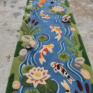 May include: A rectangular rug depicting a pond scene with koi fish, water lilies, and aquatic plants. The rug features a blue pond with colorful koi fish in shades of orange, yellow, black, and white. Green plants and pink water lilies surround the pond.