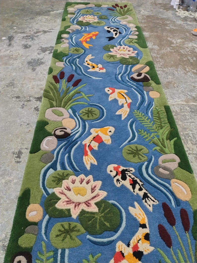 May include: A rectangular rug depicting a pond scene with koi fish, water lilies, and aquatic plants. The rug features a blue pond with colorful koi fish in shades of orange, yellow, black, and white. Green plants and pink water lilies surround the pond.