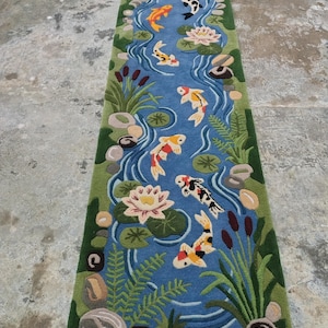 May include: A rectangular rug featuring a pond scene with koi fish, water lilies, and aquatic plants. The rug has a blue background with a green border. The koi fish are orange, white, and black.