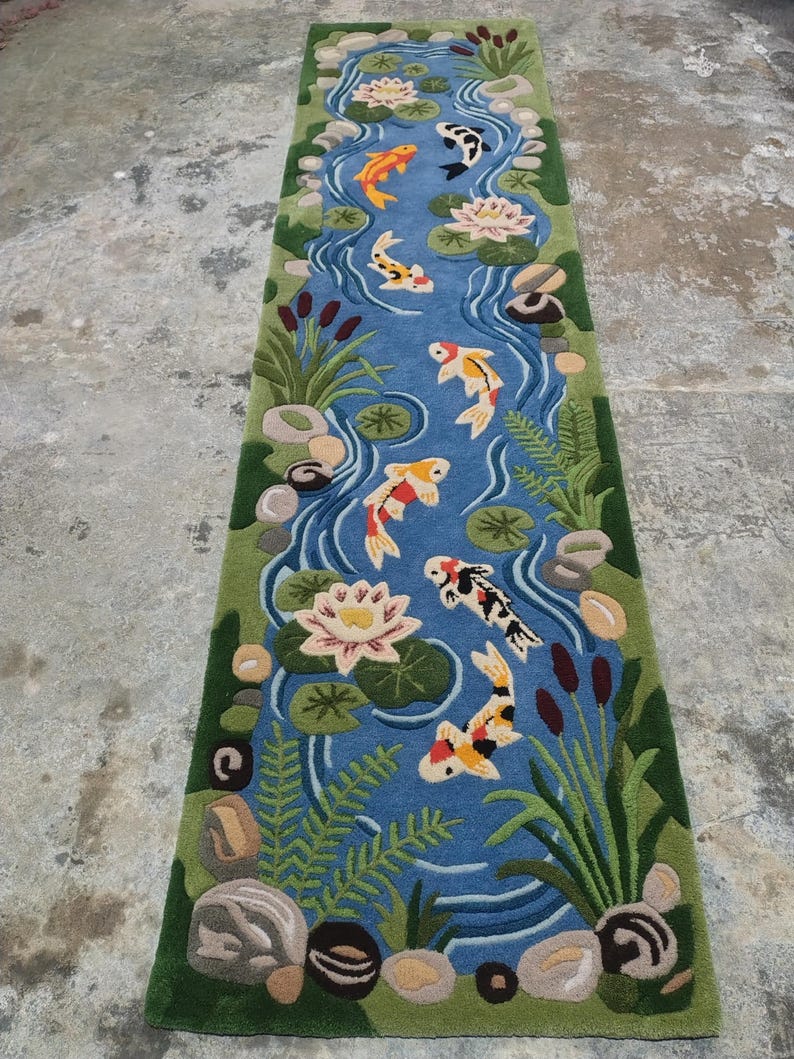 May include: A rectangular rug featuring a pond scene with koi fish, water lilies, and aquatic plants. The rug has a blue background with a green border. The koi fish are orange, white, and black.