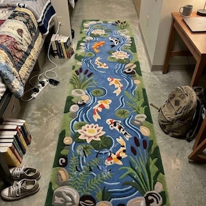May include: A rectangular rug featuring a blue river scene with koi fish, water lilies, and aquatic plants. The rug has a green border and is designed to resemble a pond. The koi fish are orange, black, and white.