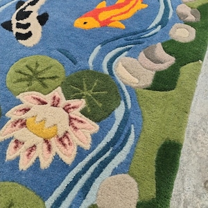 May include: A detailed rug depicting a serene pond scene. The design features two koi fish, one black and white, the other orange and yellow, swimming in blue water. Lily pads, a water lily, and stones add to the naturalistic aesthetic. Green and beige borders frame the scene.