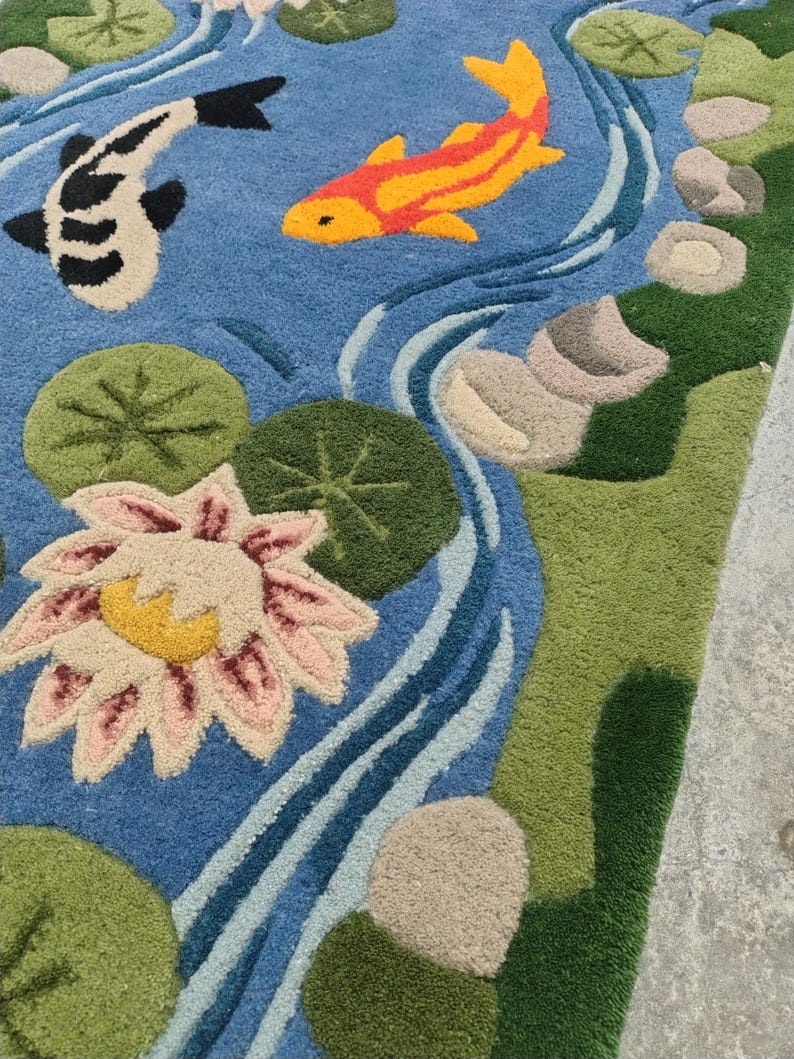 May include: A detailed rug depicting a serene pond scene. The design features two koi fish, one black and white, the other orange and yellow, swimming in blue water. Lily pads, a water lily, and stones add to the naturalistic aesthetic. Green and beige borders frame the scene.