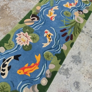 May include: A rectangular rug featuring a blue river scene with colorful koi fish, lily pads, and aquatic plants. The rug has a green border and is designed with a detailed, hand-tufted appearance. The koi fish are orange, yellow, black, and white.